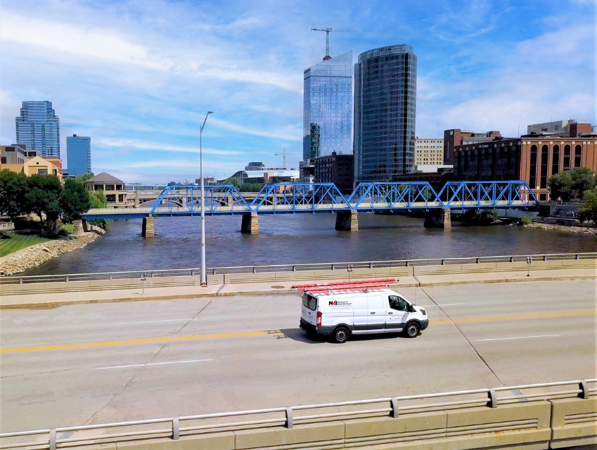 An NAI Wisinski of West Michigan service van with a red ladder on top drives across a bridge in downtown Grand Rapids. The Grand River and the iconic blue pedestrian bridge are visible in the background, along with modern high-rise buildings.